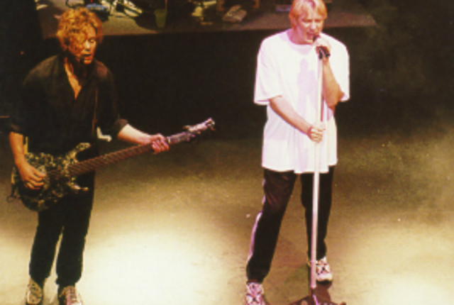 Two men on stage with guitars and microphones.