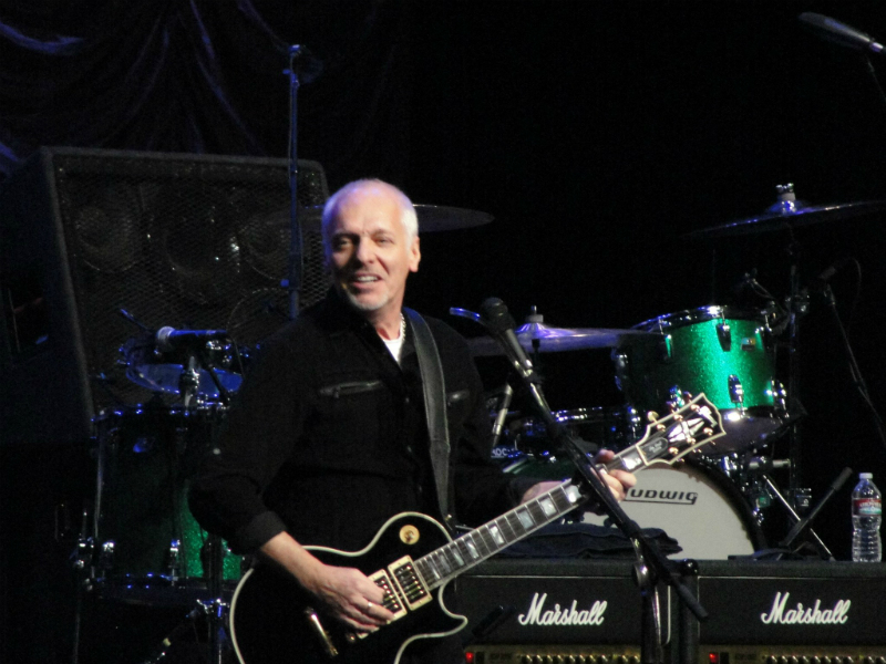 A man with a black guitar in front of a stage.